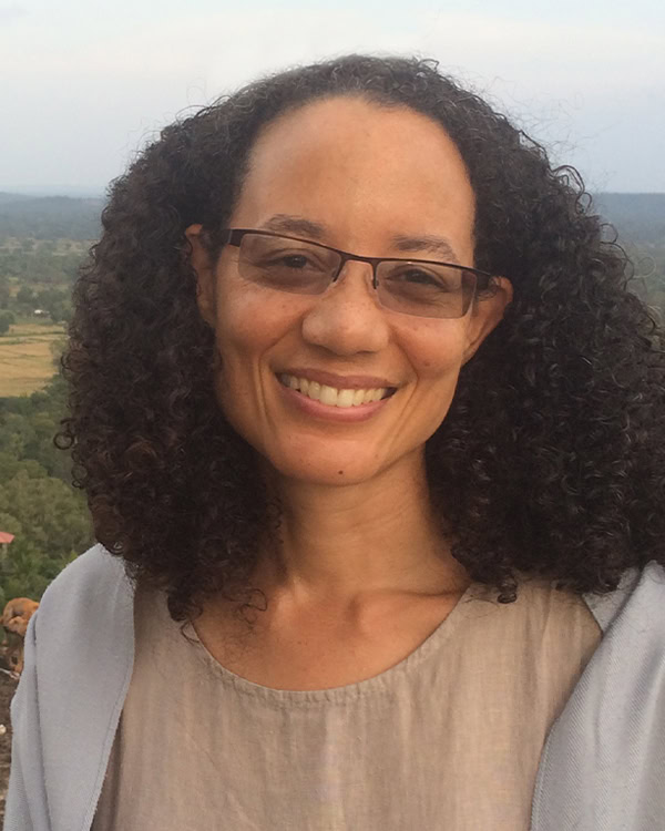 A Woman With Curly Dark Hair And Glasses Smiles At The Camera. She Is Wearing A Light Beige Top And A Light Blue Shawl, With A Natural Landscape Of Trees And Fields In The Background.