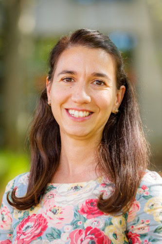 A Woman With Long Brown Hair, Wearing A Floral-Patterned Top, Smiles At The Camera While Standing Outdoors With A Blurred Green Background.