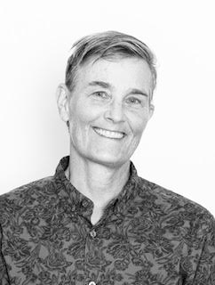 Black And White Portrait Of A Person With Short, Light Hair, Smiling And Wearing A Patterned Button-Up Shirt, Posed Against A Plain Background.